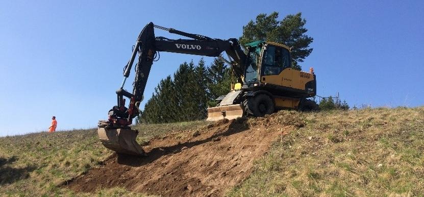 Grävmaskin gräver fram sand i en gräsbevuxen slänt.